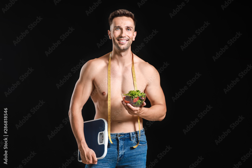 Handsome muscular man with measuring tape, salad and scales on dark background. Weight loss concept