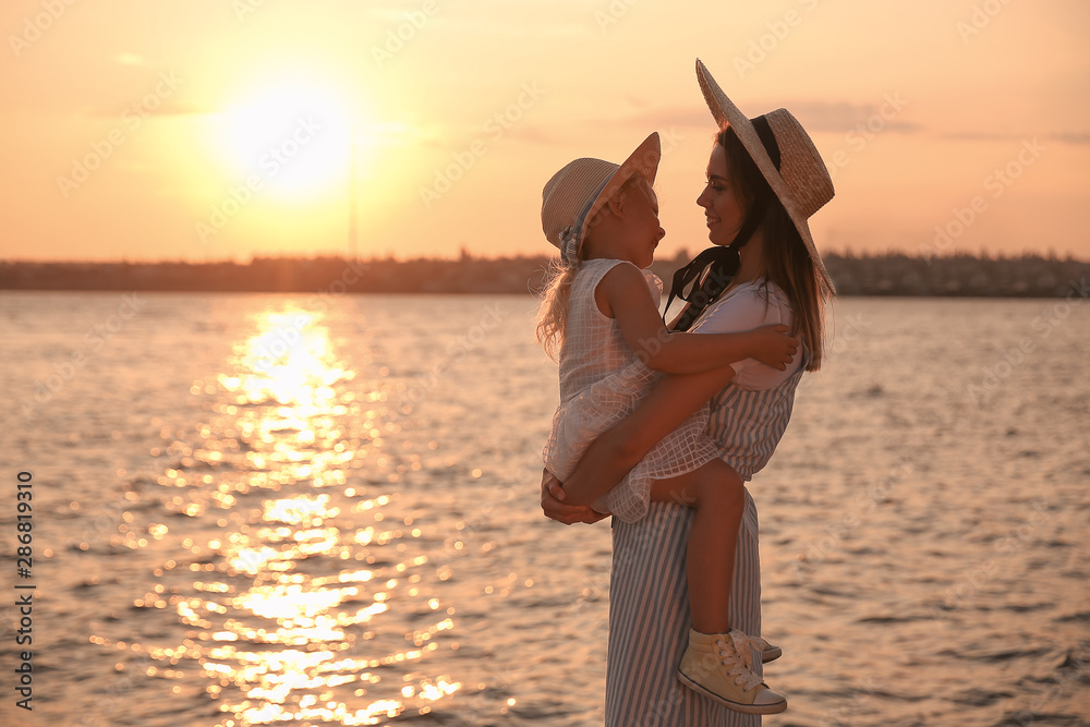 Happy mother and her little daughter near river at sunset