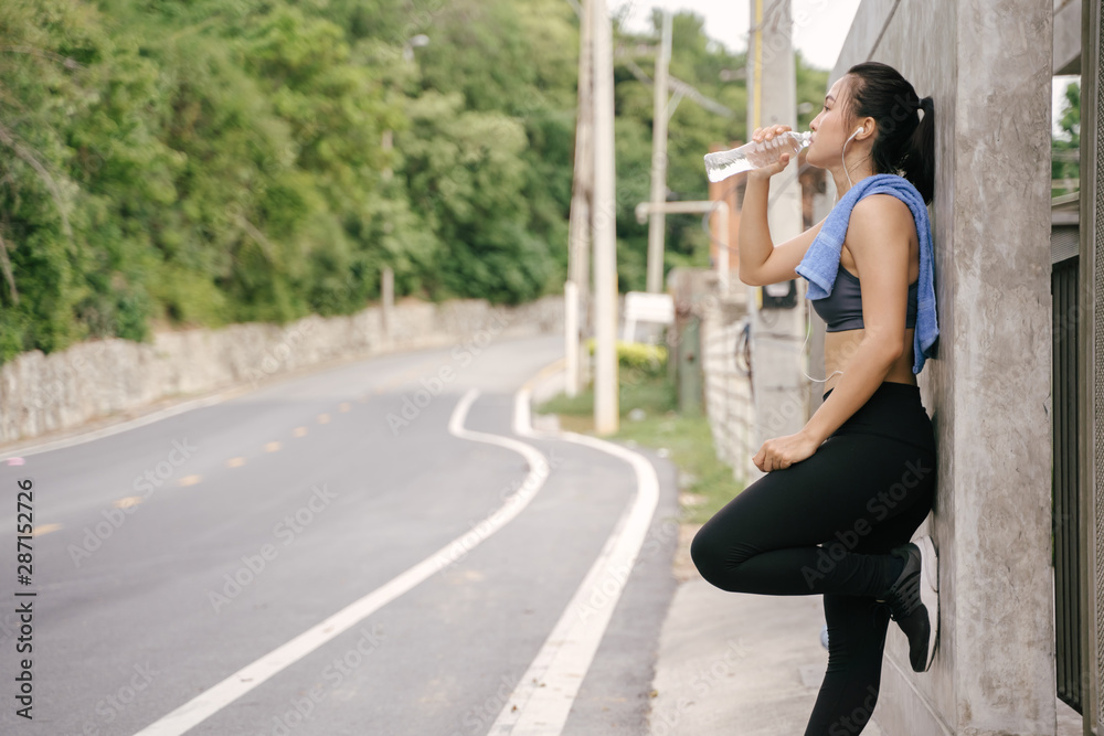 Young Asian woman runner resting after workout running and drinking water in street road.