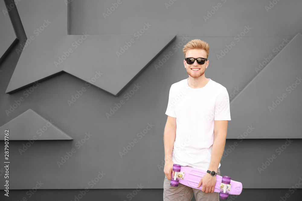 Man in stylish t-shirt and with skateboard near grey wall