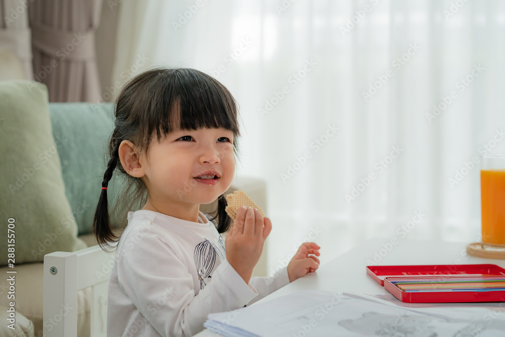 快乐的亚洲蹒跚学步的女孩，在客厅里对着她画画时，一边吃着威化饼干，一边露出灿烂的笑容