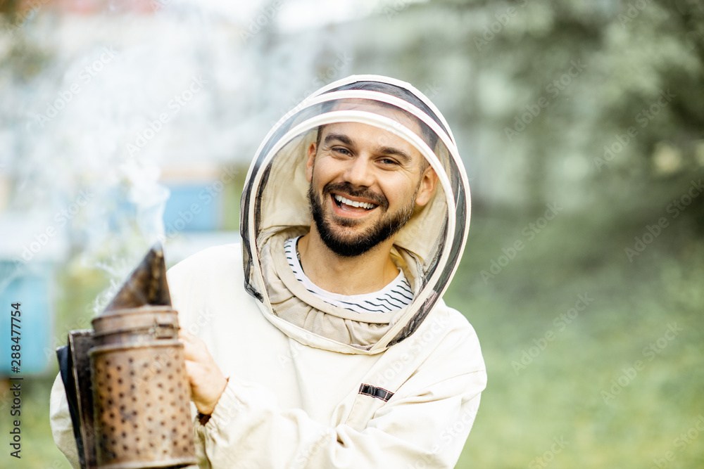 Portrait of a cheerful beekeeper in protective uniform with bee smoker on the apiary