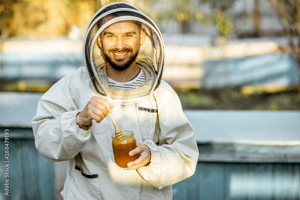 Portrait of a handsome beekeper in protective uniform standing with honey, tasting fresh product on 