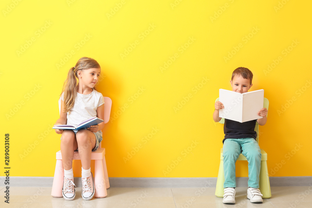 Little children with books sitting on chairs near color wall