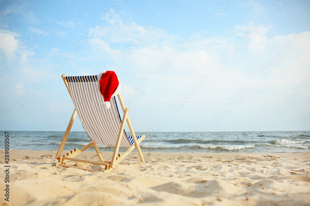 Beach chair with Santa Claus hat on sea coast. Christmas vacation