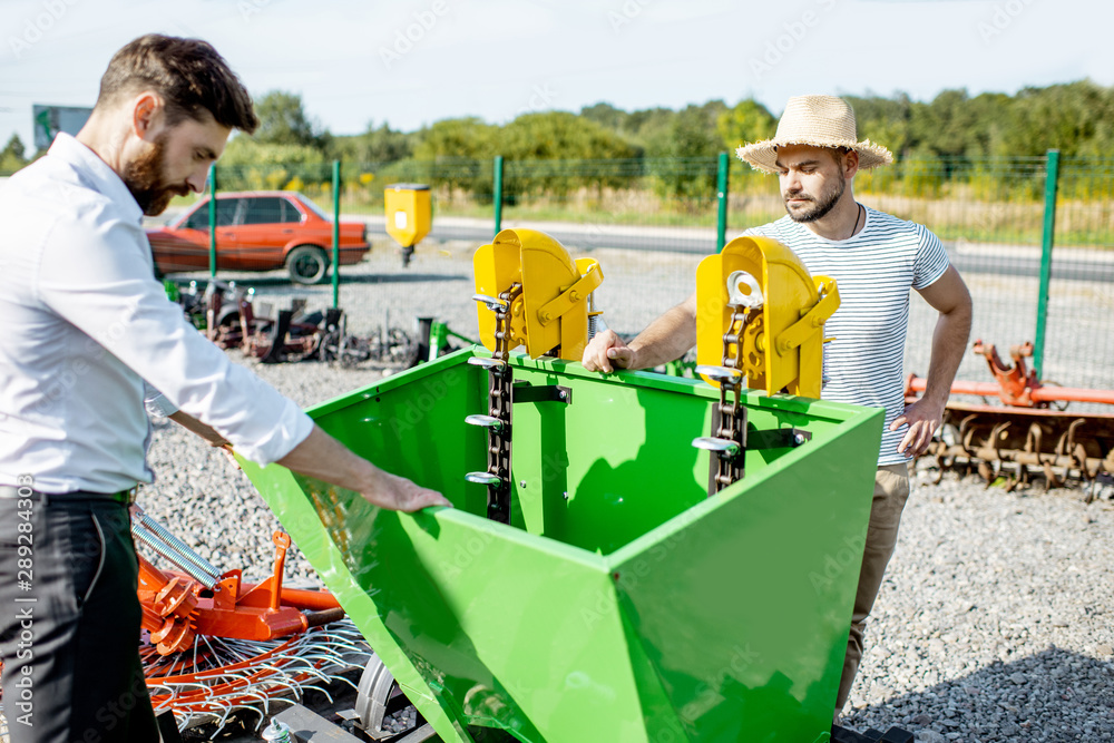 Young agronomist with salesman at the open ground of the shop with agricultural machinery, buying a 