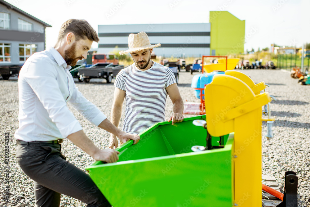 Young agronomist with salesman at the open ground of the shop with agricultural machinery, buying a 