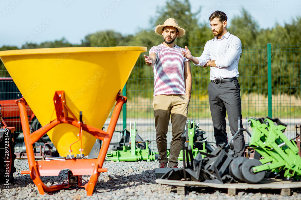 Young agronomist with salesman at the open ground of the shop with agricultural machinery, buying a 