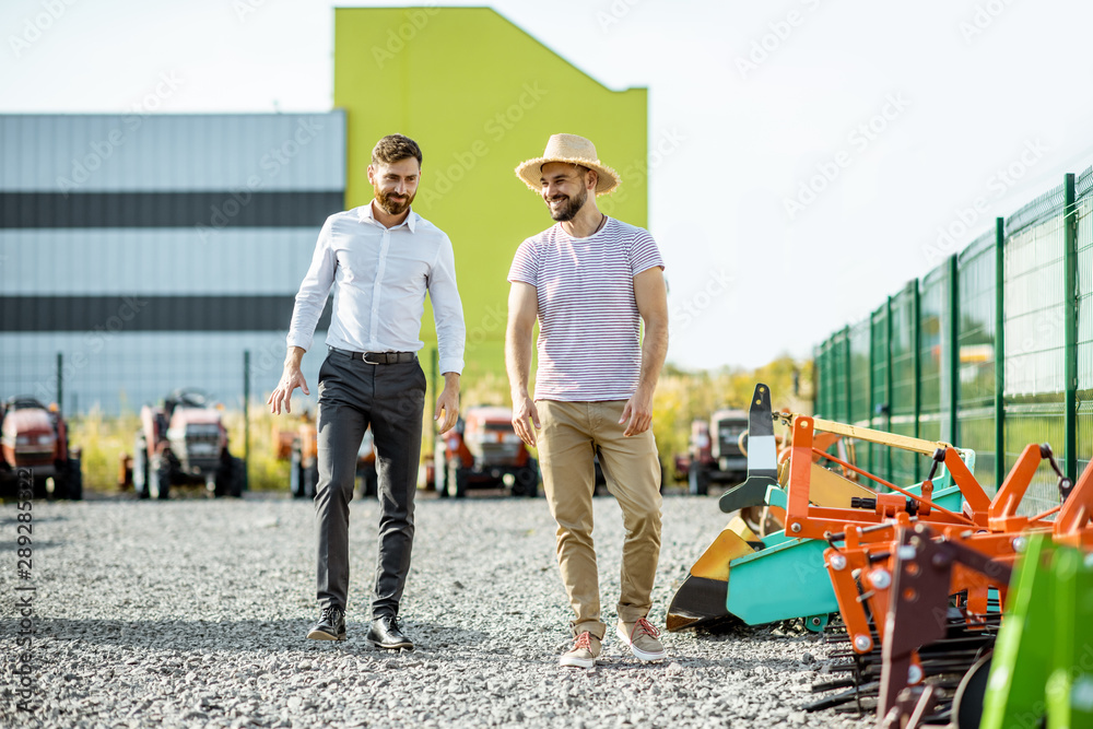 Young agronomist walking with salesman at the open ground of the shop with agricultural machinery, c