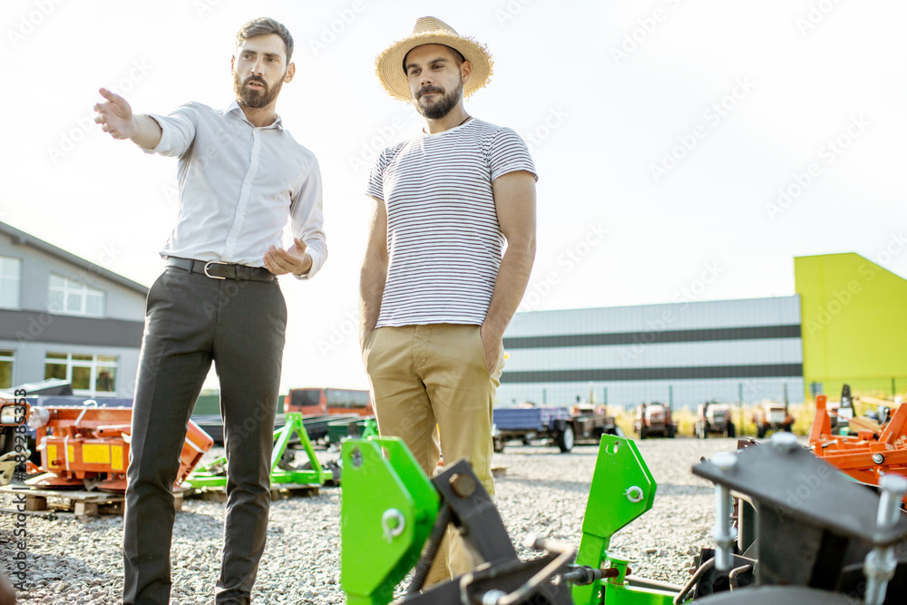 Young agronomist walking with salesman at the open ground of the shop with agricultural machinery, c