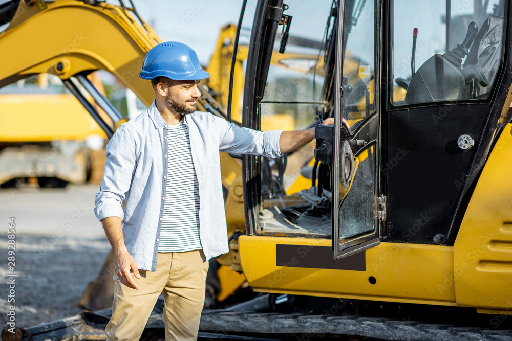 Portrait of a handsome builder standing near the escavator on the open ground of the shop with heavy