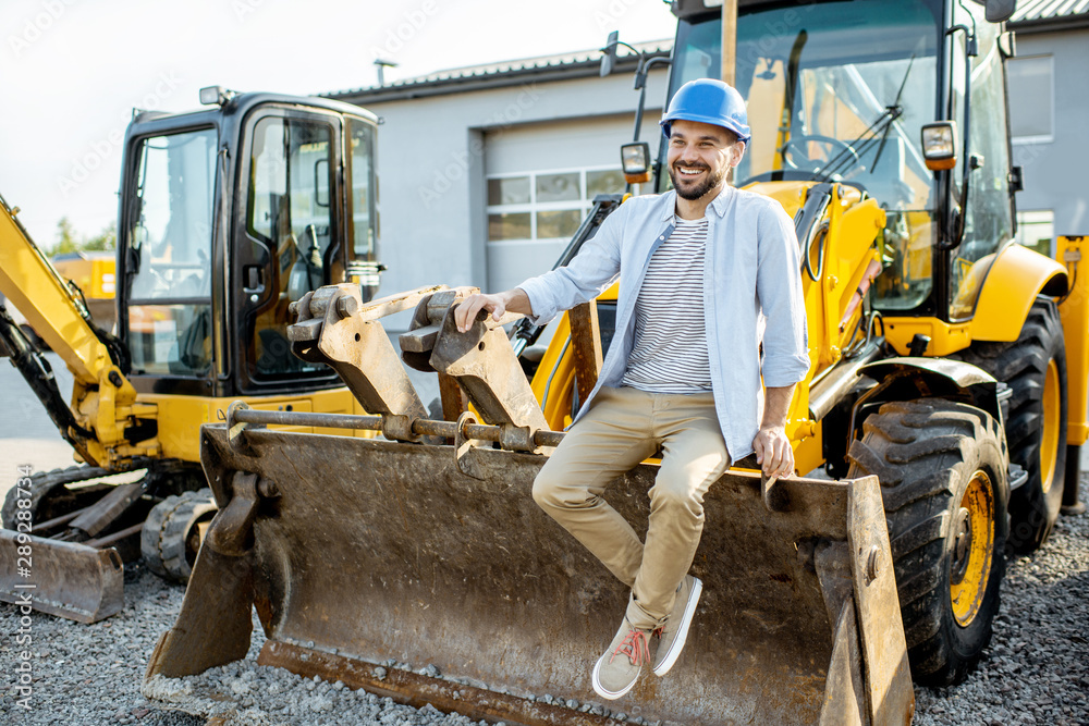 Portrait of a handsome builder sitting on a bulldozer scoop on the open ground of the shop with heav