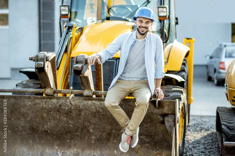 Portrait of a handsome builder sitting on a bulldozer scoop on the open ground of the shop with heav