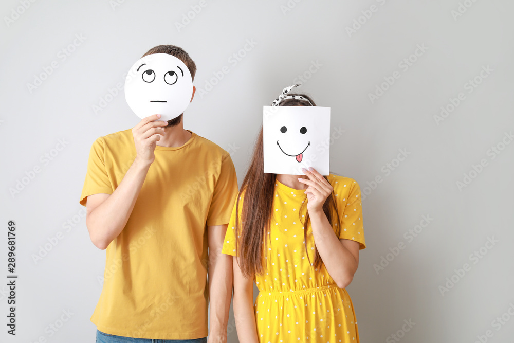 Couple hiding faces behind sheets of paper with drawn emoticons on grey background