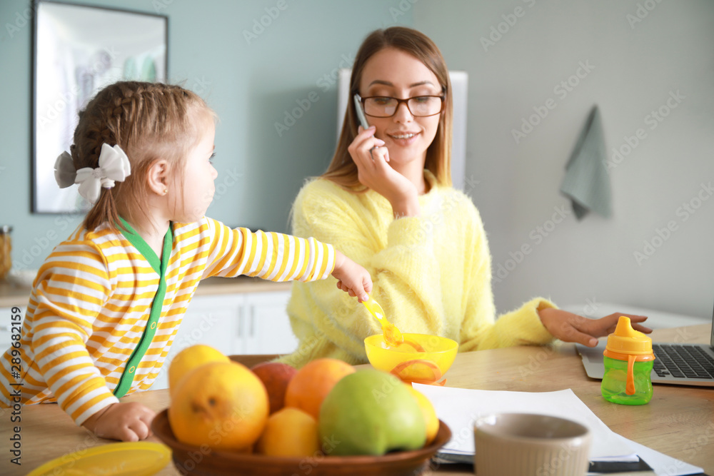 Little daughter with working mother in kitchen at home