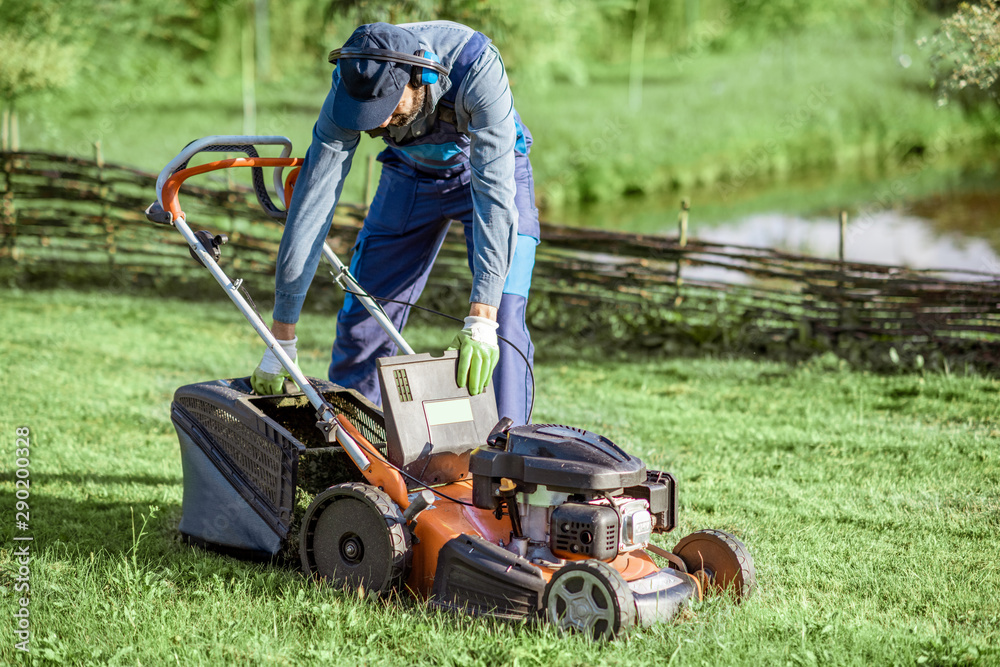 Professional gardener in protective workwear getting grass basket from the lawn mower while cutting 