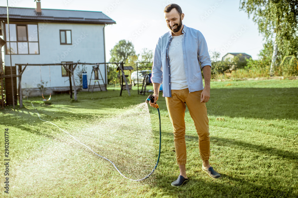 Handsome man watering green lawn, sprinkling water on the grass during a sunny morning on the backya