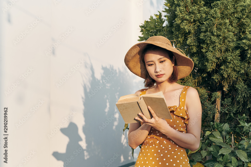 Young woman reading a book standing lean against trunk tree in summer park outdoor.