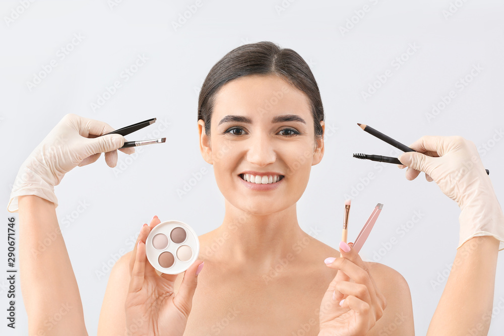 Young woman with beautiful eyebrows and hands of cosmetologist holding tools against light backgroun