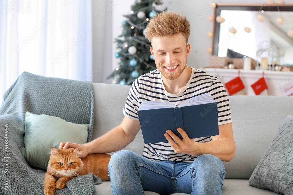 Young man with cute cat reading book at home on Christmas eve