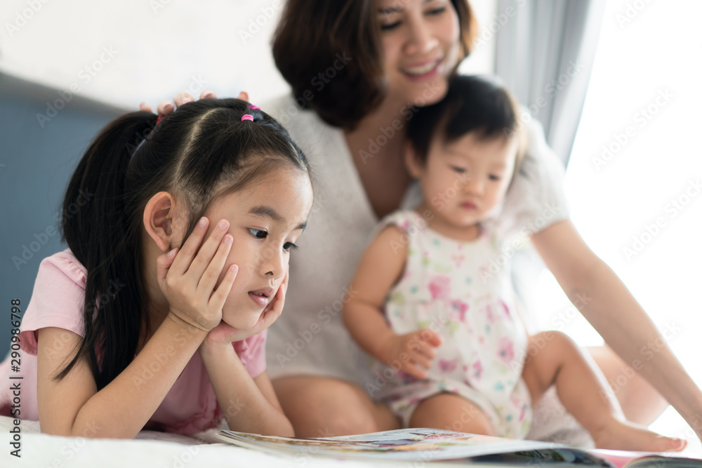 Beautiful Asian mother reading fairly tale story book with her children on the bed. Mom pointing the