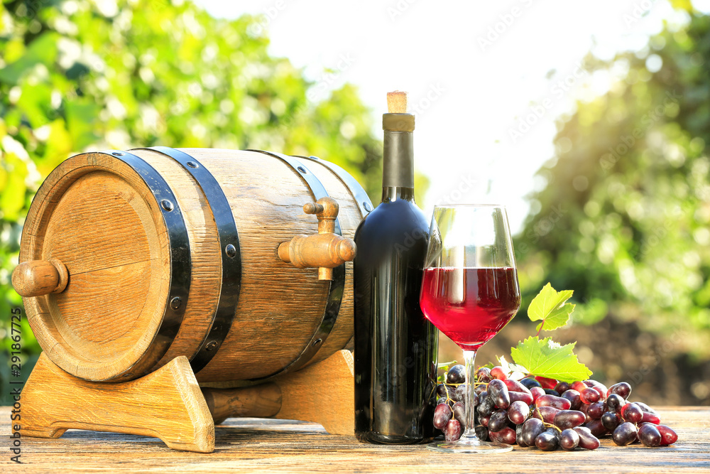 Glass and bottle of red wine with fresh grapes and barrel on wooden table in vineyard