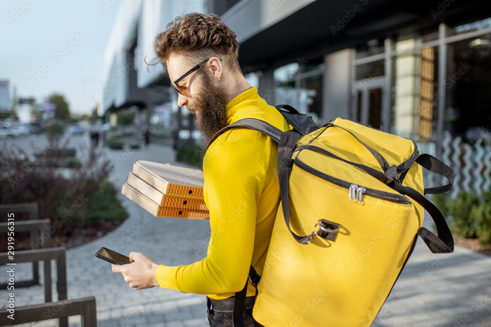 Young male courier in yellow sweatshirt delivering pizza, standing with thermal backpack and smart p