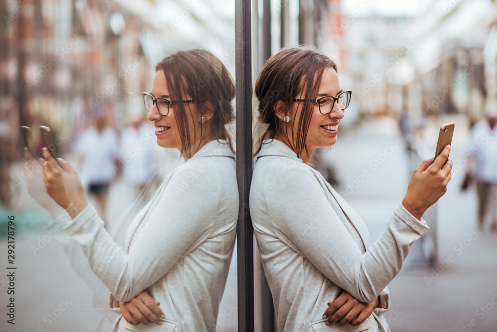 Portrait of a beautiful young woman leaning on a glass wall and using smart phone in the city.