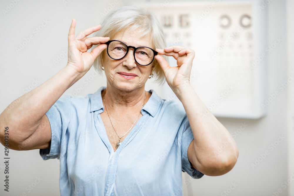 Portrait of a happy senior woman wearing eyeglasses in front of eye chart in ophthalmology office. C