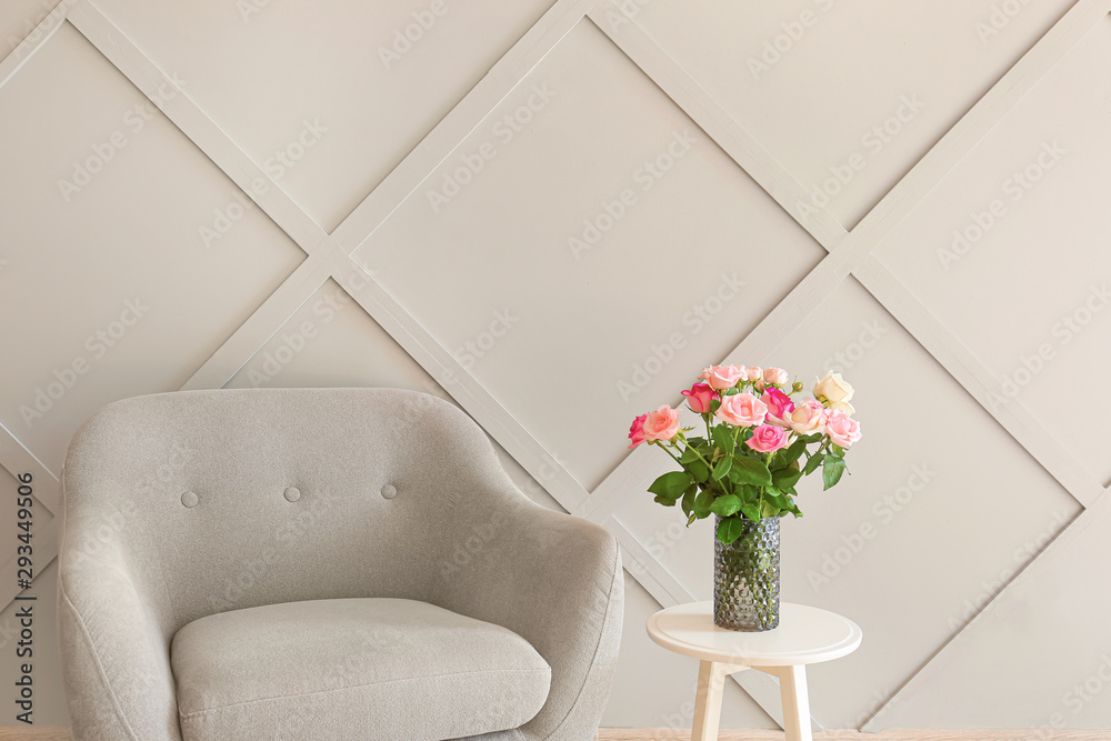 Beautiful rose flowers in vase on table and armchair near grey wall in room