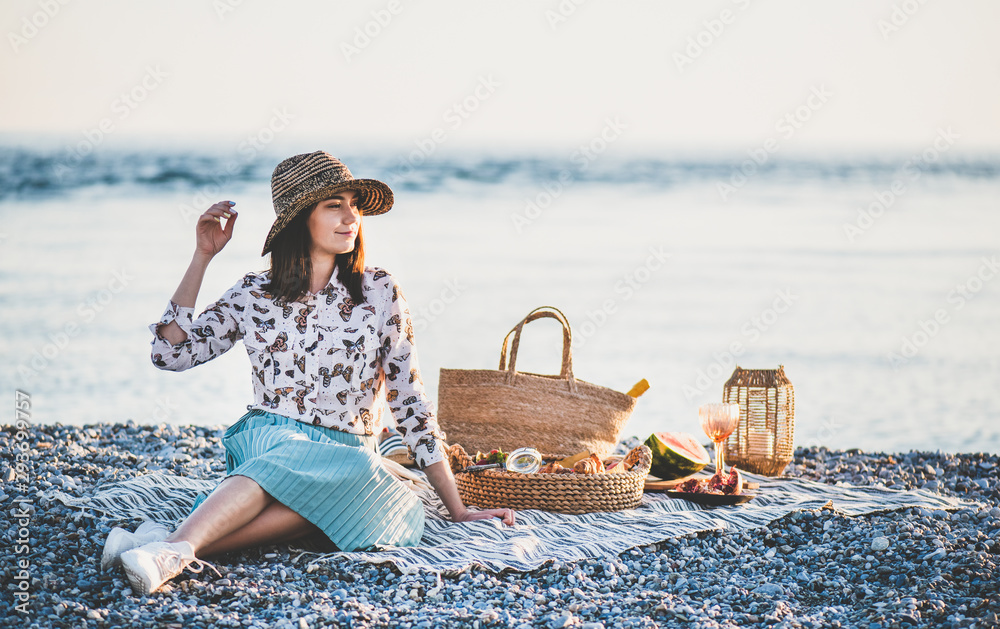 日落时分的夏季海滩野餐。戴帽子的年轻女子坐在毯子上，周末户外野餐