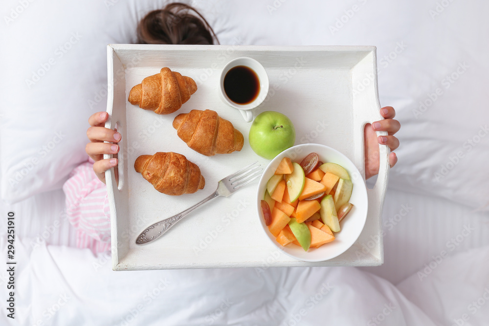 Young woman holding tray with tasty breakfast while lying in bed