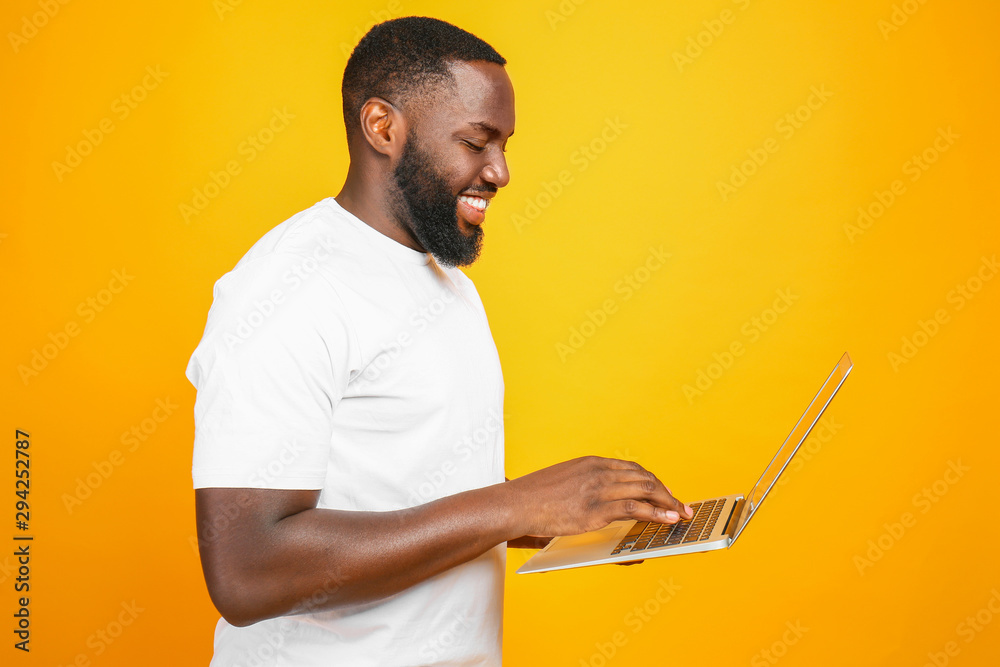 Handsome African-American man with laptop on color background