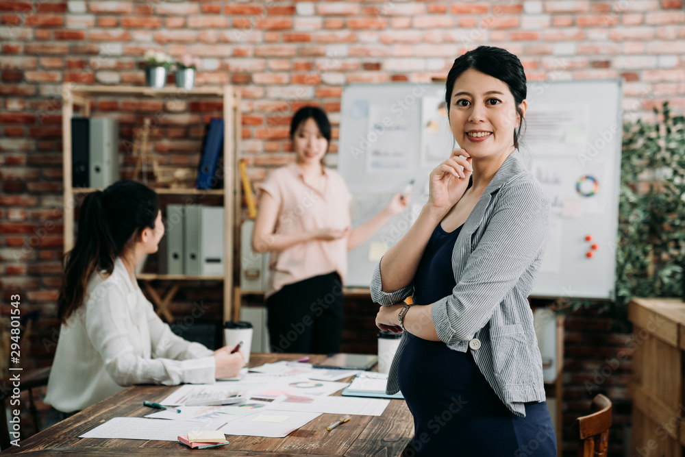 Confident young Asian pregnant businesswoman standing with folded arms smiling at camera in boardroo