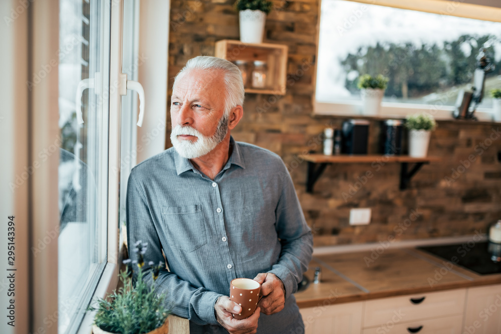 Portrait of a senior man standing in the kitchen and looking through window, holding a cup of coffee