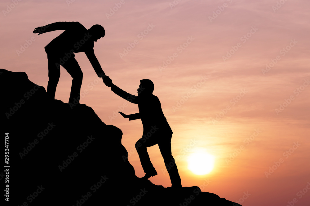 Silhouette of people helping each other hike up a mountain at sunset background. Teamwork, success a