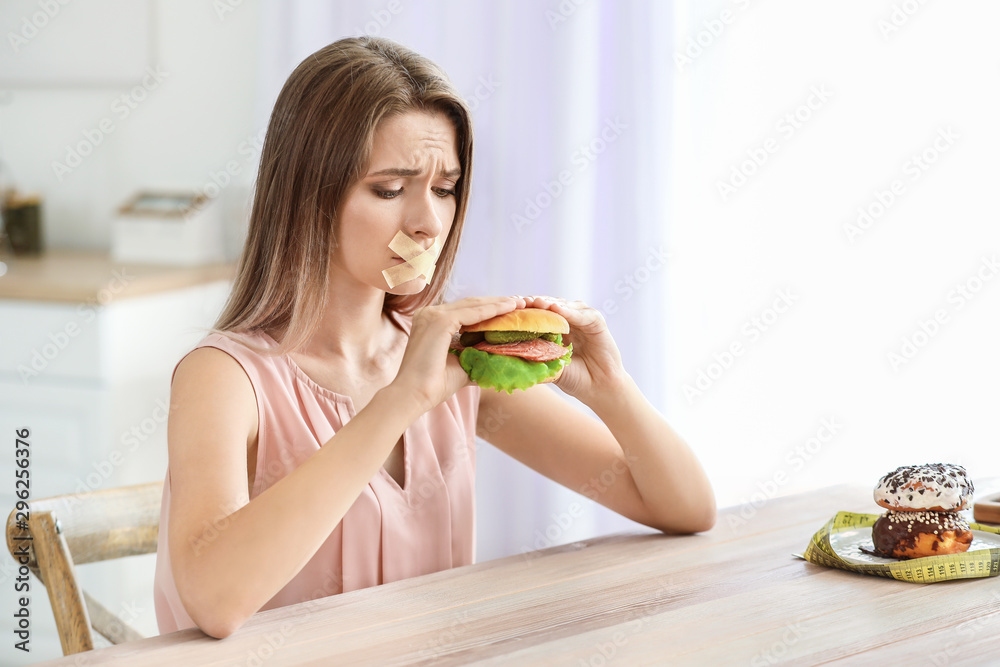 Sad woman with taped mouth and with tasty burger in kitchen. Diet concept