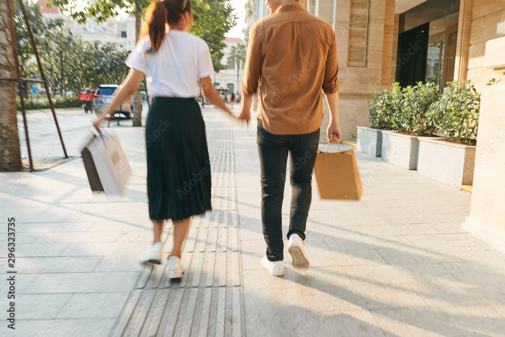 Lower body section of a young tourist couple walking by store windows and holding paper shopping bag