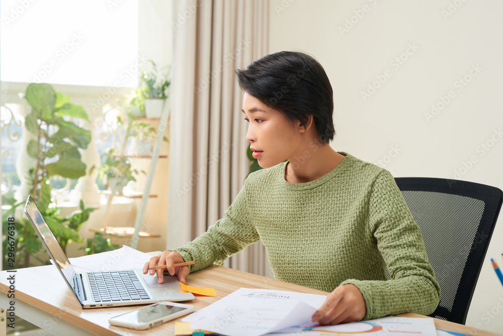 Concentrated Asian woman sit at table working on laptop making notes in notebook at home office