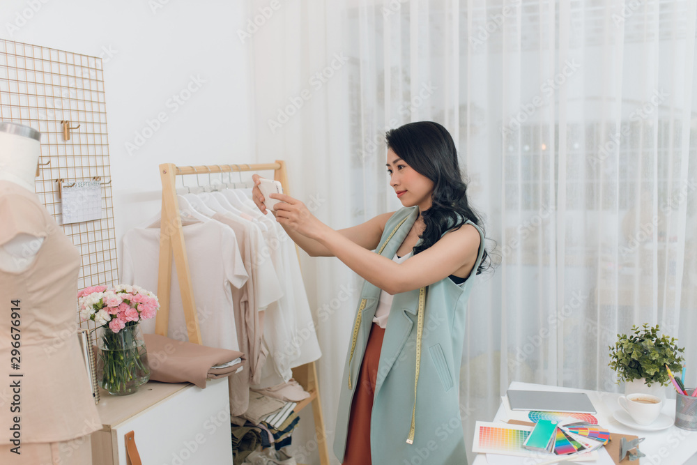 Beautiful young asian fashion designer taking photo of work using smartphone in dressmaking studio.