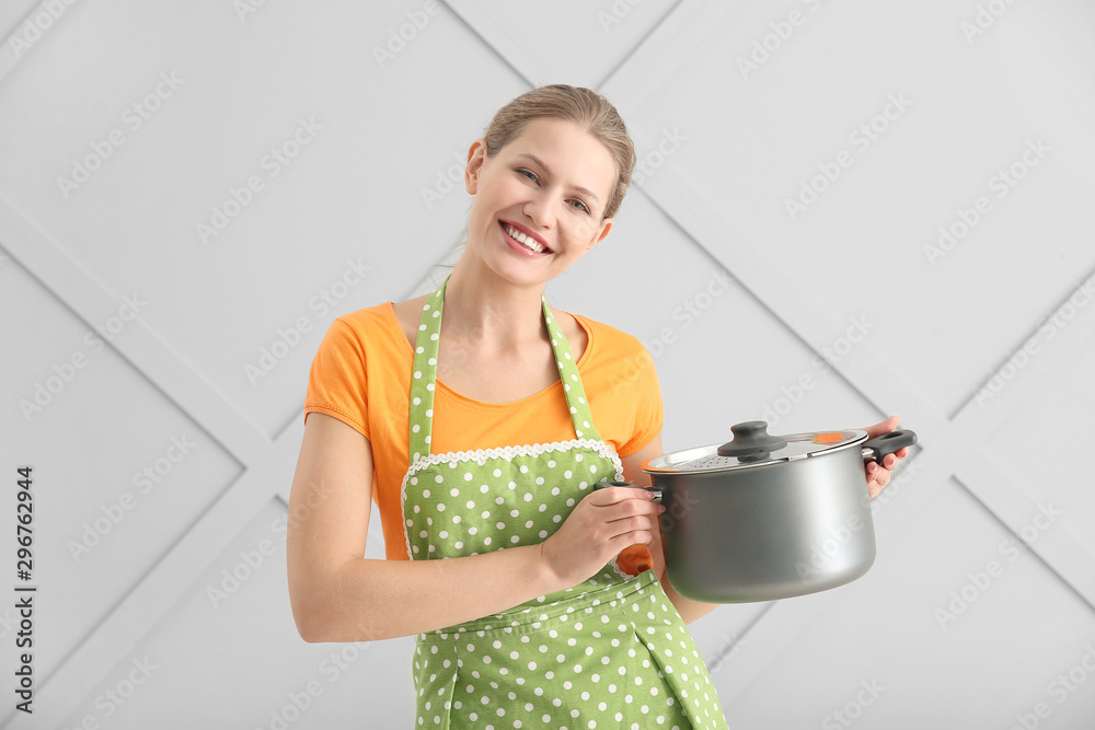 Beautiful young woman in apron and with saucepan on light background