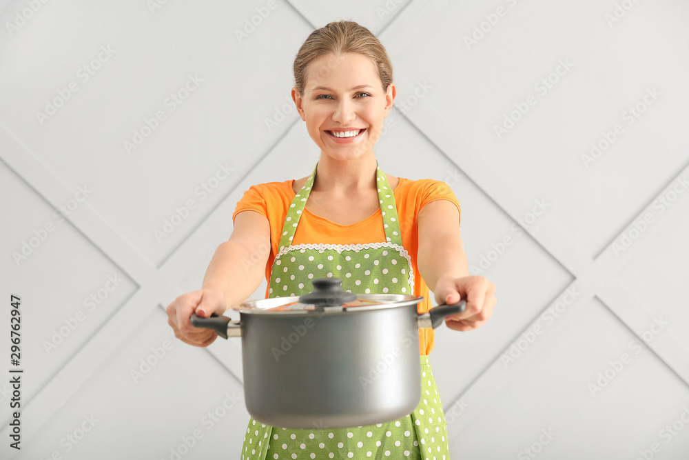 Beautiful young woman in apron and with saucepan on light background