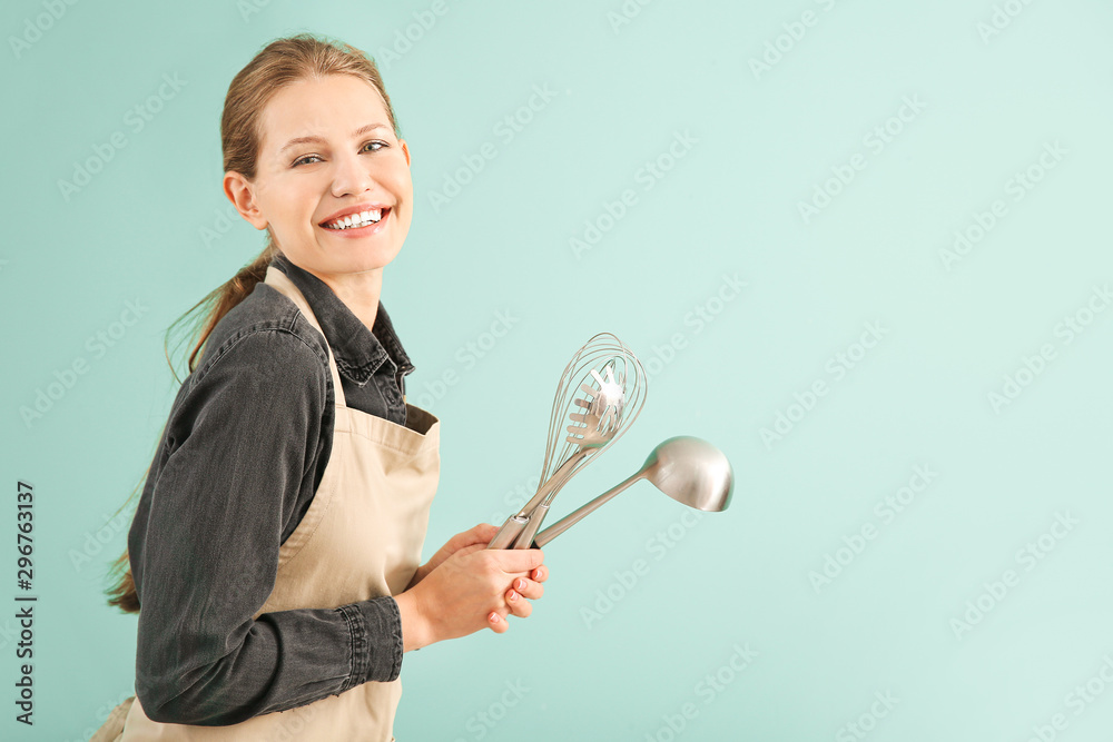 Beautiful young woman in apron and with kitchenware on color background