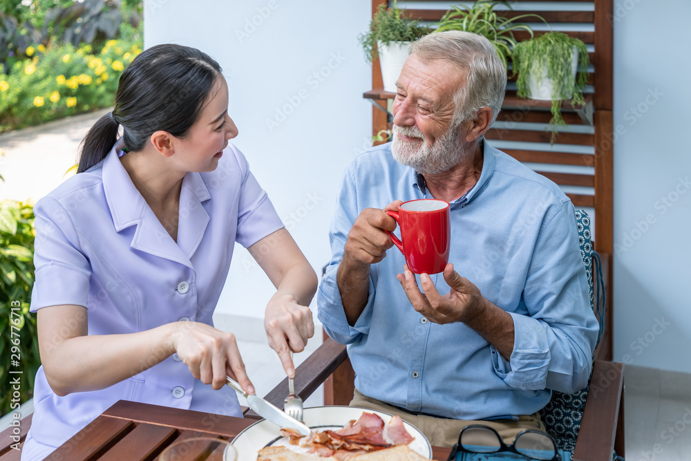 Nurse assist elderly senior man to eat breakfast and drink coffee with mug in hand at nursing home