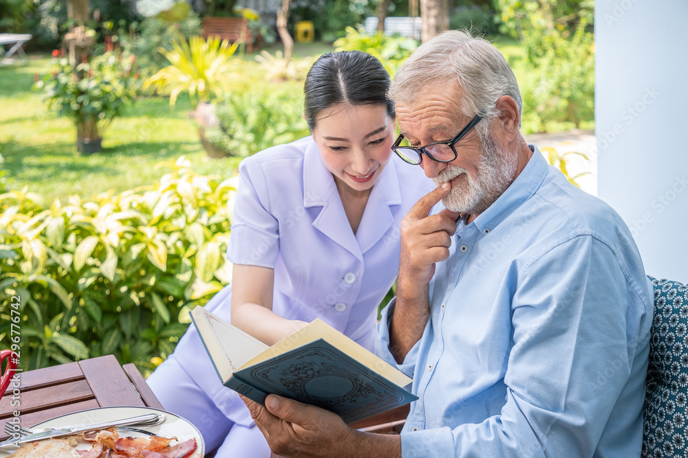 Senior elderly man reading book with nurse during breakfast in garden at nursing home