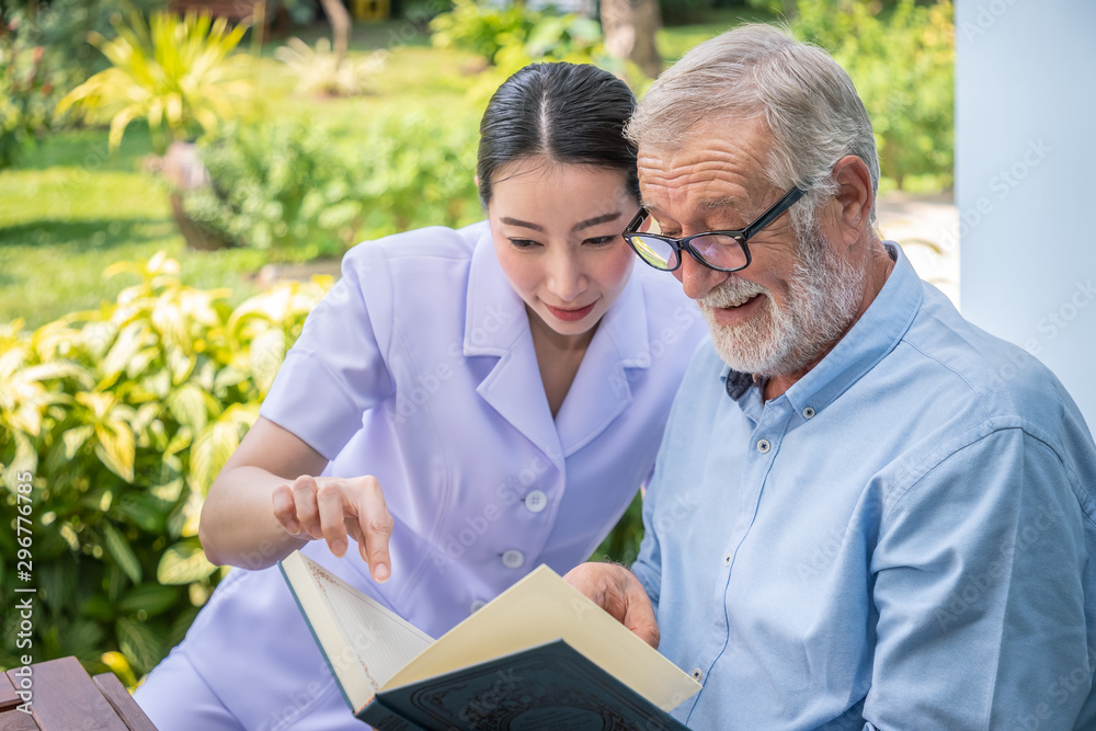 Senior elderly man reading book with nurse during breakfast in garden at nursing home