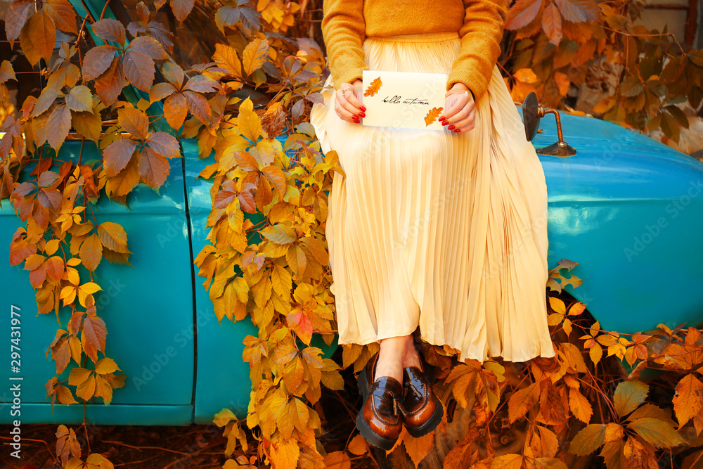 Beautiful young woman with greeting card sitting on car outdoors on autumn day