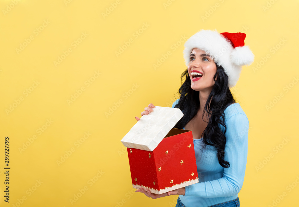 Young woman with santa hat opening a Christmas gift box on a yellow background