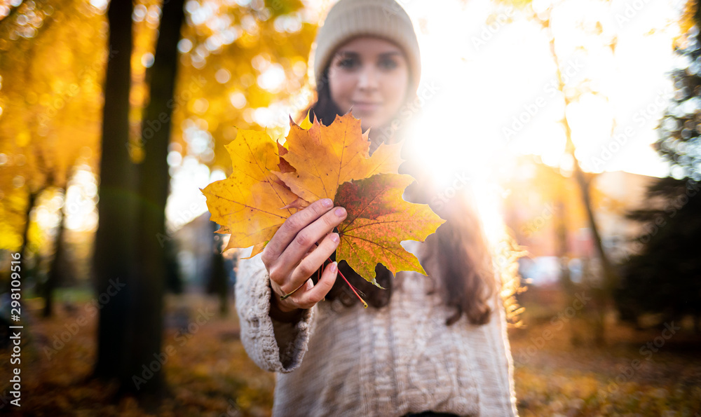 Outdoor lifestyle portrait of young woman with warm hat holding colorful autumn leaves and smiling