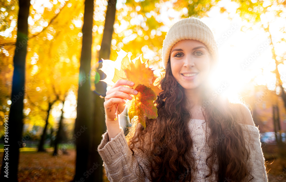 Outdoor lifestyle portrait of young woman with warm hat holding colorful autumn leaves and smiling